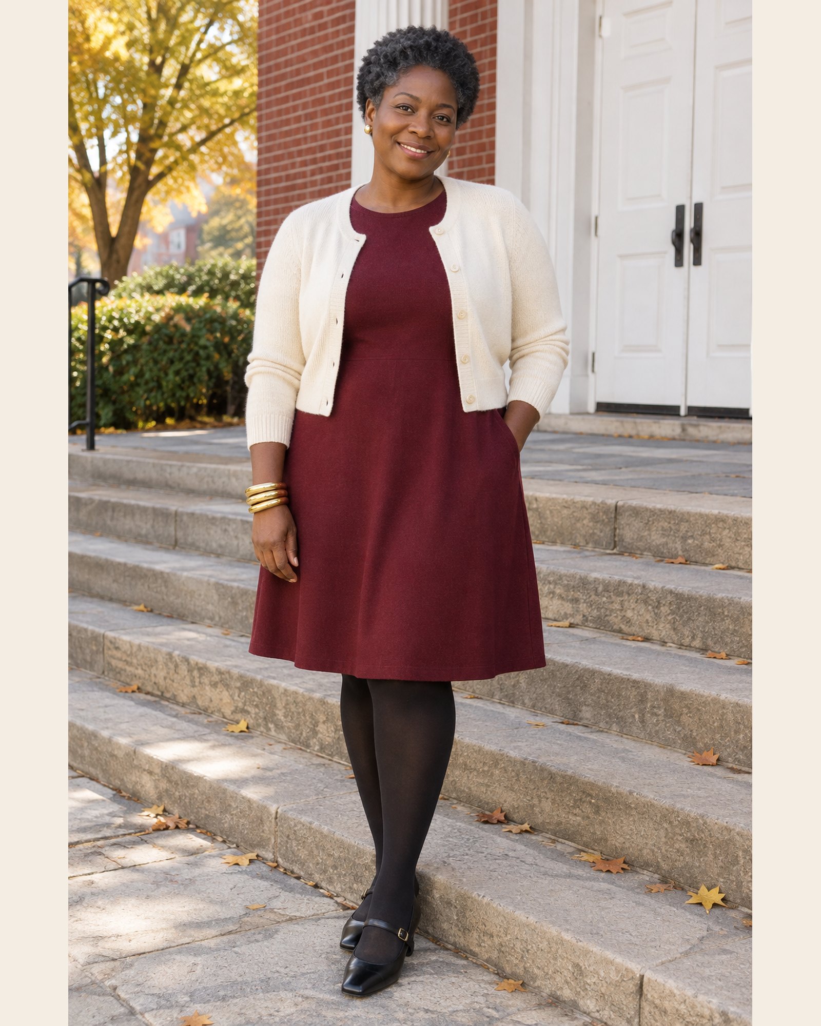 AI-generated image: a fictional model wearing a burgundy knee-length knit dress, a cropped cream cardigan, black opaque tights, black Mary Jane flats and a stack of gold bangles, on stone steps in front of a brick building in autumn.